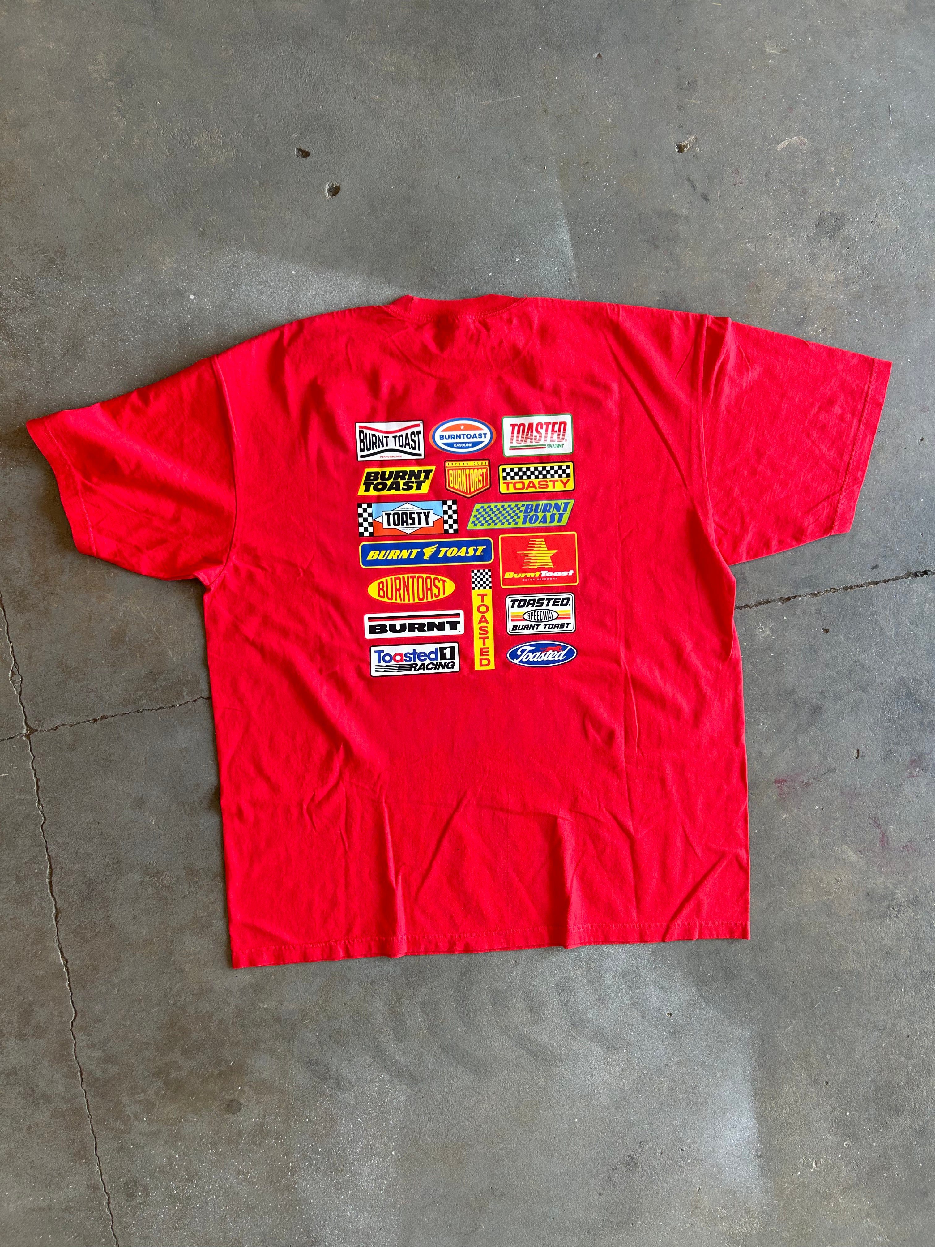 A red short-sleeve t-shirt laid flat on a gray concrete floor, featuring a large graphic print on the back with various colorful racing sponsor logos.