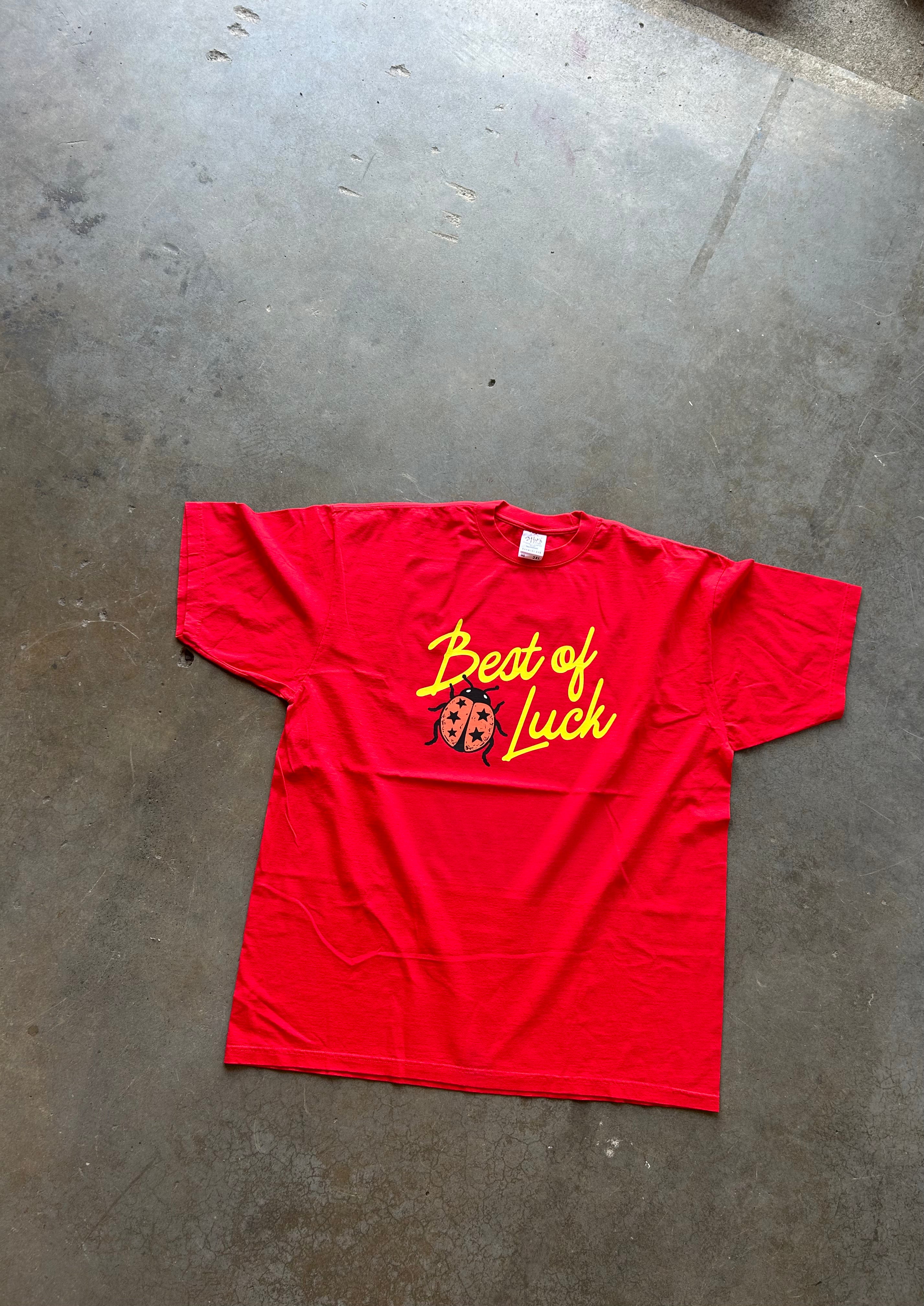 A bright red short-sleeve t-shirt laid flat on a gray concrete floor. The front of the shirt features a graphic with yellow cursive text reading 'Beat of Luck' and a small ladybug illustration.