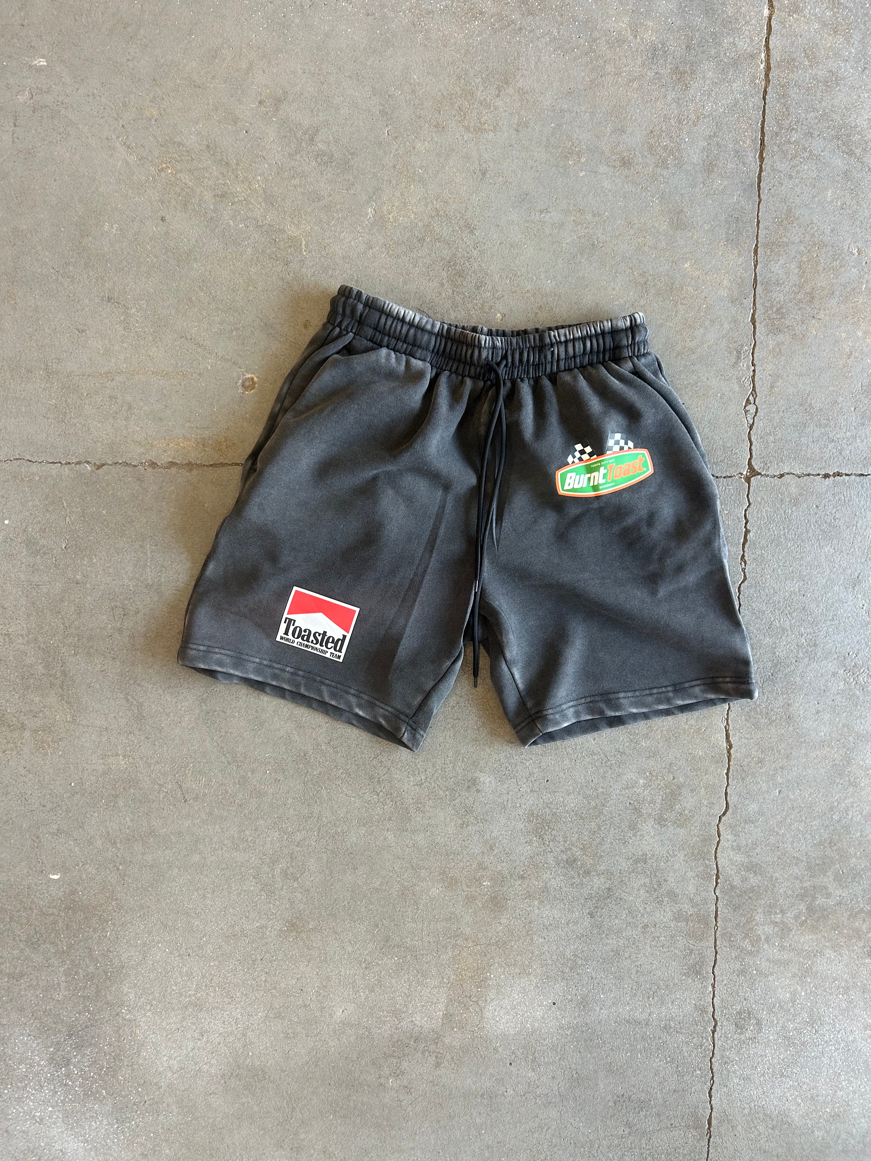 Black athletic shorts with an elastic waistband, featuring a red and white 'Torsion' patch on the left leg and a green and white 'Buckeye' patch on the right leg, laid flat on a concrete floor