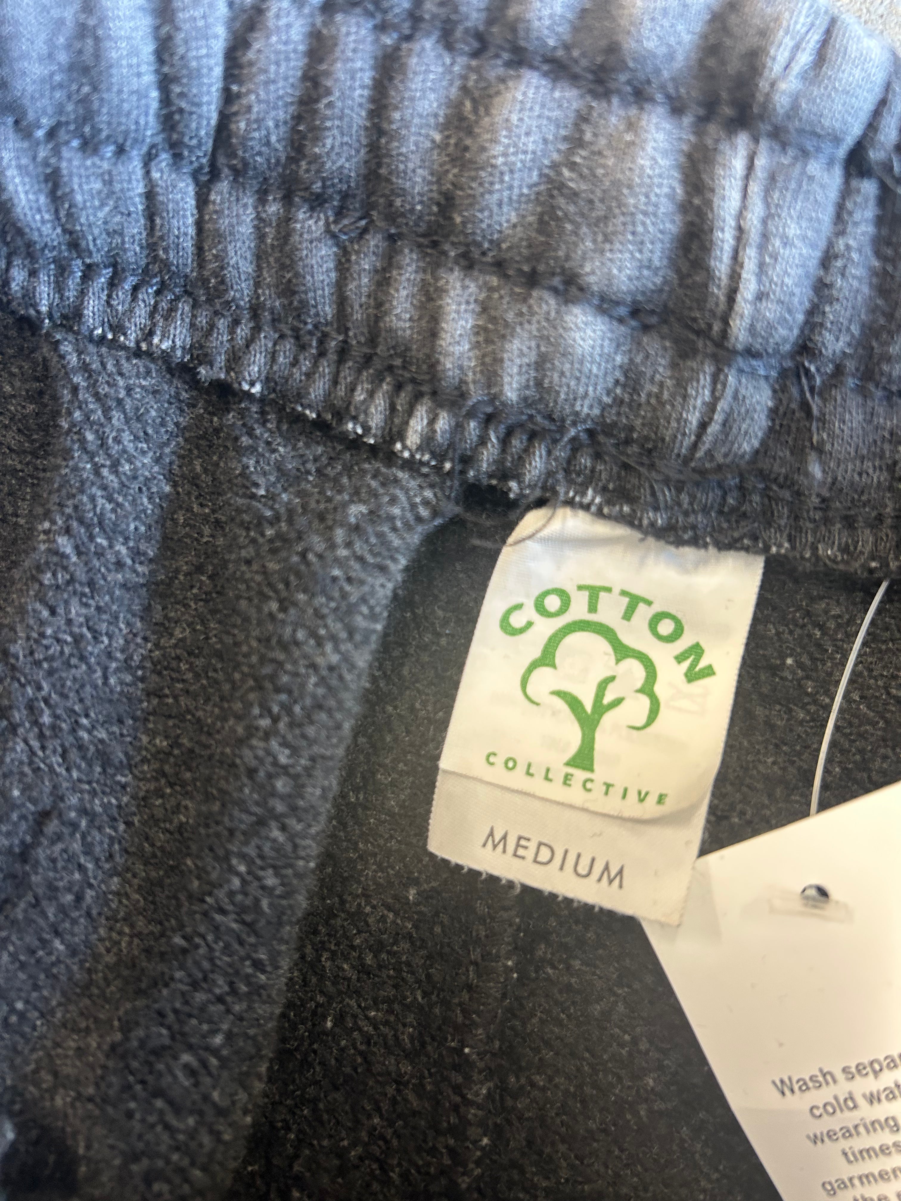 Close-up of a gray heathered fabric garment with a sewn-on label reading COTTON COLLECTIVE and MEDIUM, plus a white care tag attached