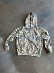 A camouflage pullover hoodie with a woodland leaf and bark pattern in shades of green, brown, and beige, laid flat on a gray concrete floor.
