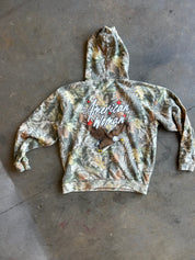 A camouflage hooded sweatshirt laid flat on a concrete floor, featuring a large graphic on the back with the words 'American Woman' and an eagle illustration.