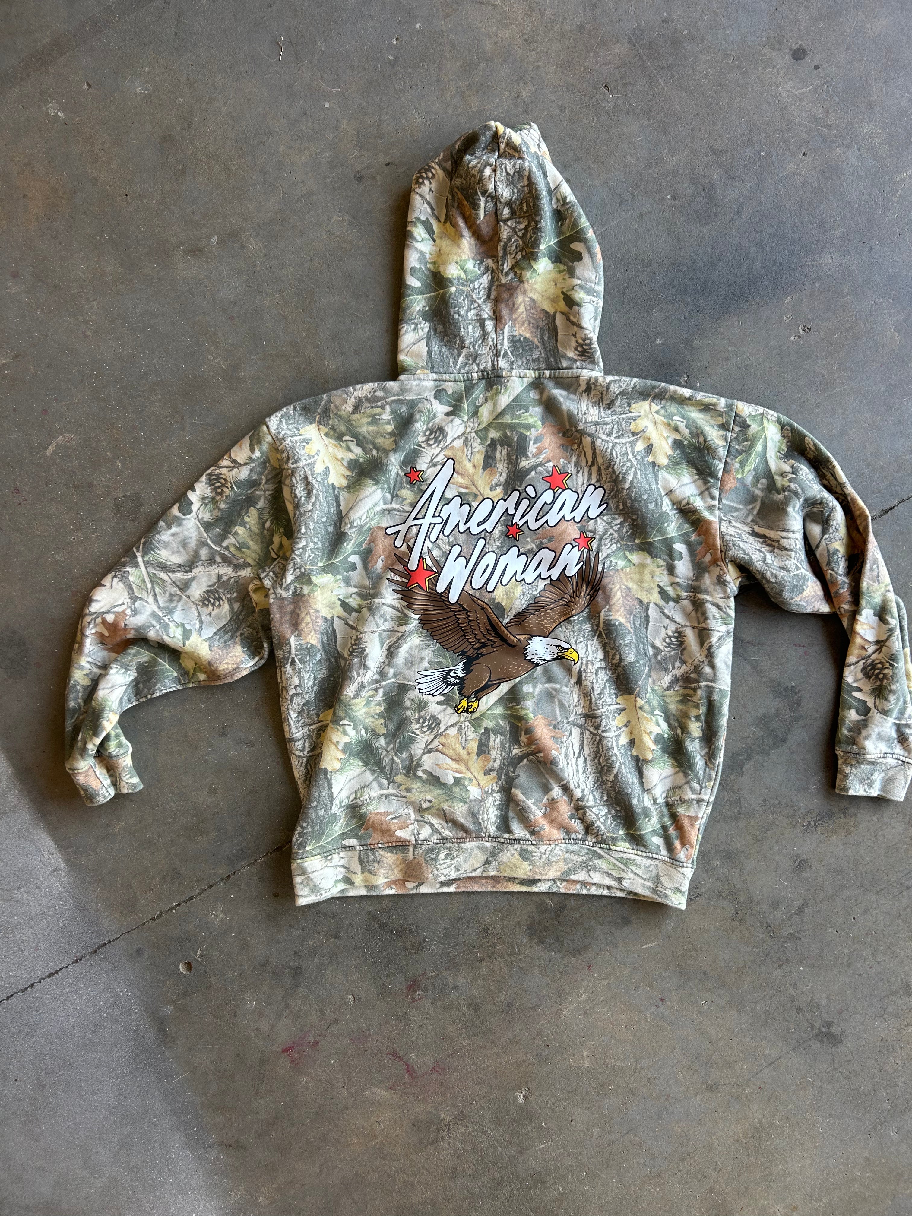 A camouflage hooded sweatshirt laid flat on a concrete floor, featuring a large graphic on the back with the words 'American Woman' and an eagle illustration.