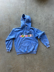 A blue washed denim-style hoodie with a front kangaroo pocket and a colorful graphic reading 'Will Death We Do Art' across the chest, laid flat on a textured gray concrete surface.
