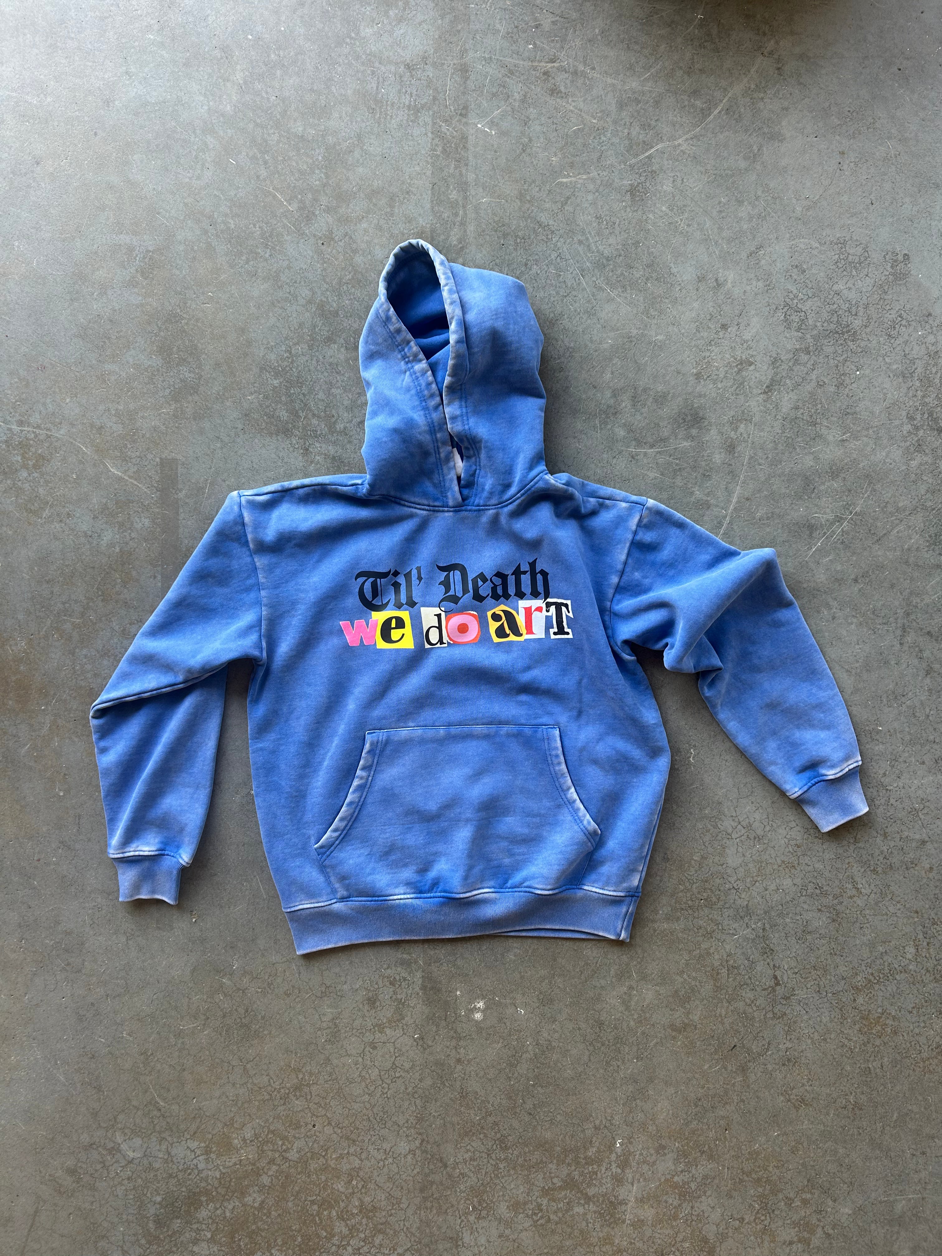 A blue washed denim-style hoodie with a front kangaroo pocket and a colorful graphic reading 'Will Death We Do Art' across the chest, laid flat on a textured gray concrete surface.