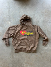 A brown leopard-print hoodie with a graphic on the chest featuring a red heart and the text 'Fragile' in yellow, laid flat on a gray concrete floor.