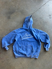 A blue denim-style hooded sweatshirt laid flat on a concrete floor, with white script text reading 'Chaos Coordinator' across the chest