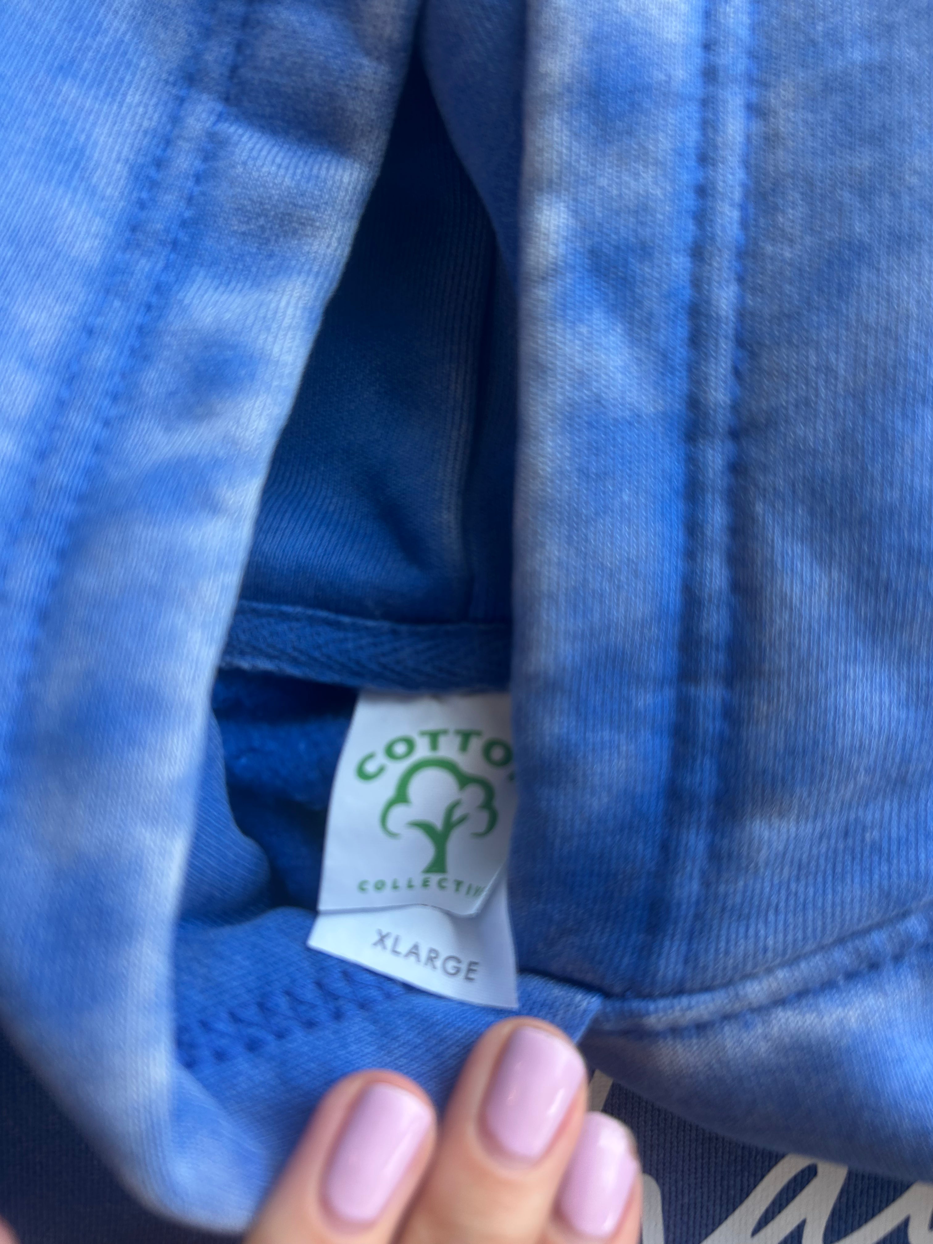 Close-up of a blue fabric garment showing a sewn-in label that reads 'COTTON COLLECTION' with a cotton tree logo and 'XLARGE' below, with a hand with light pink nails holding the fabric open.