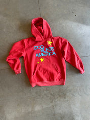 A red pullover hoodie with 'GOD BLESS AMERICA' printed in blue and yellow on the front, laid flat on a gray concrete floor.