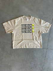 A beige short-sleeve t-shirt with 'WEIRD' printed in black letters next to three yellow alien heads, laid flat on a gray concrete floor.