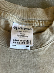 Close-up of the inside neck of a beige cotton t-shirt showing the brand label 'SHAKA WEAR' and 'DROP SHOULDER GARMENT DYE'.