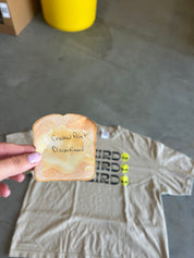 A hand holds a toasted slice of bread with handwritten text reading 'Cracked Print Discontinued' in front of a beige graphic T-shirt laid on a concrete floor. The shirt has a black graphic and three small yellow alien faces.