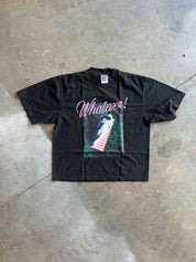 A black short-sleeve graphic t-shirt laid flat on a concrete floor. The shirt features the word 'Whatever!' in pink cursive script above a graphic