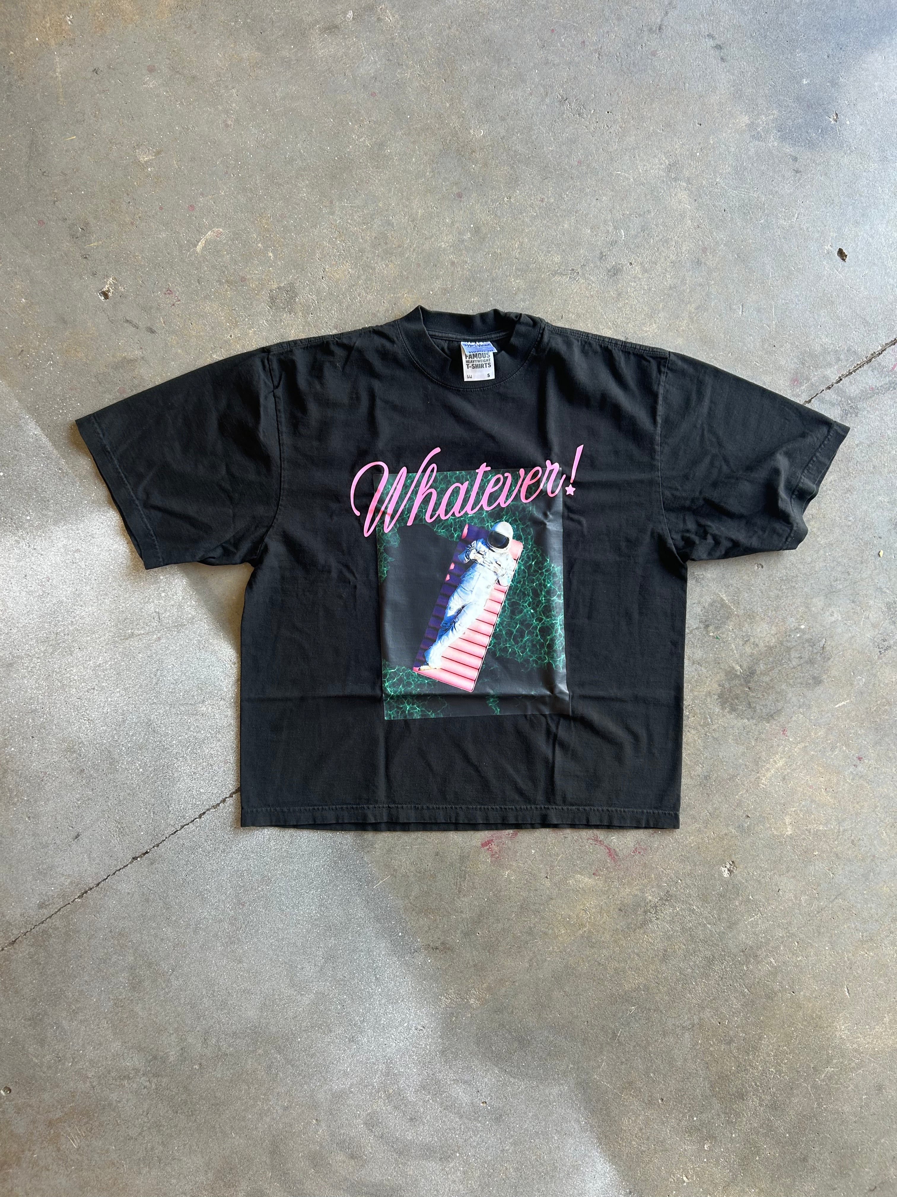A black short-sleeve graphic t-shirt laid flat on a concrete floor. The shirt features the word 'Whatever!' in pink cursive script above a graphic