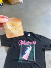 A hand holds a toasted bread slice with the words 'Crooked Print' written on it above a black graphic t-shirt on a concrete floor. The t-shirt features a pink 'Whatever!' script and a colorful print on the front.