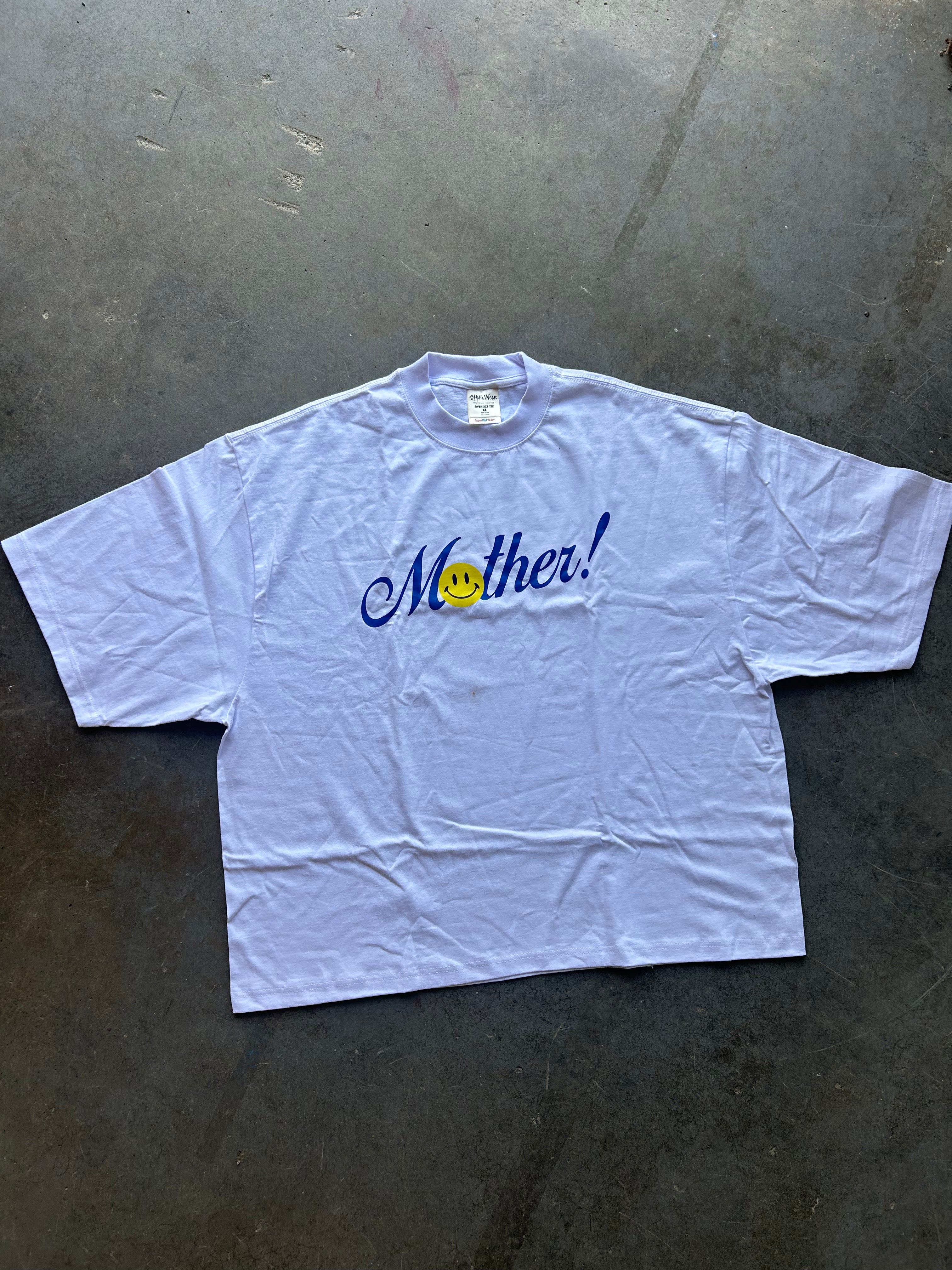 A light blue short-sleeve t-shirt laid flat on a dark concrete floor, featuring the word 'Mother!' in blue cursive with a yellow smiley face graphic.