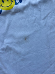 A close-up of wrinkled light blue fabric with a small dark stain in the center and part of a yellow smiley face graphic with blue lettering visible at the top left.