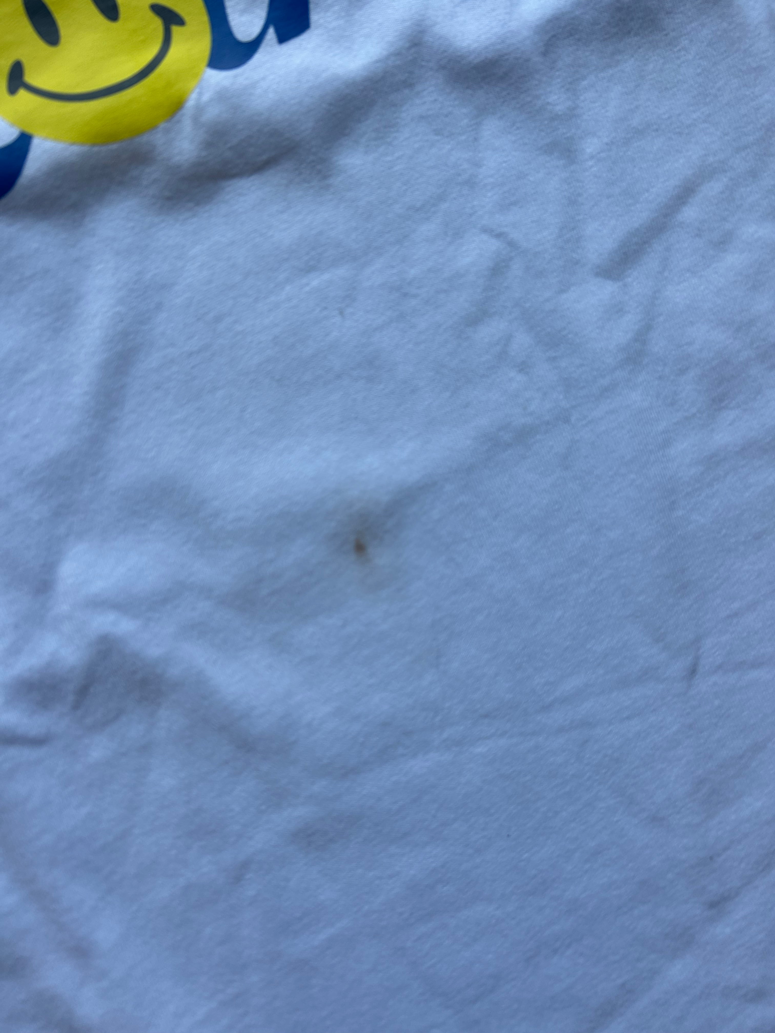 A close-up of wrinkled light blue fabric with a small dark stain in the center and part of a yellow smiley face graphic with blue lettering visible at the top left.