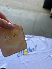 A hand with lavender-painted nails holds a stained, water-damaged copy of a toast-shaped sticker or decal over a white t-shirt that says 'Motha!'.
