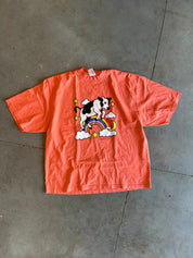 A coral orange short-sleeve t-shirt laid flat on a gray concrete floor, featuring a graphic of a black and white cow riding a rainbow with white clouds and yellow stars.