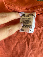 A close-up of an orange crewneck t-shirt showing the inside neck label which reads 'BHAKA WEAR' and 'Top Shoulder Garment Dye'. A hand is holding the label in place.