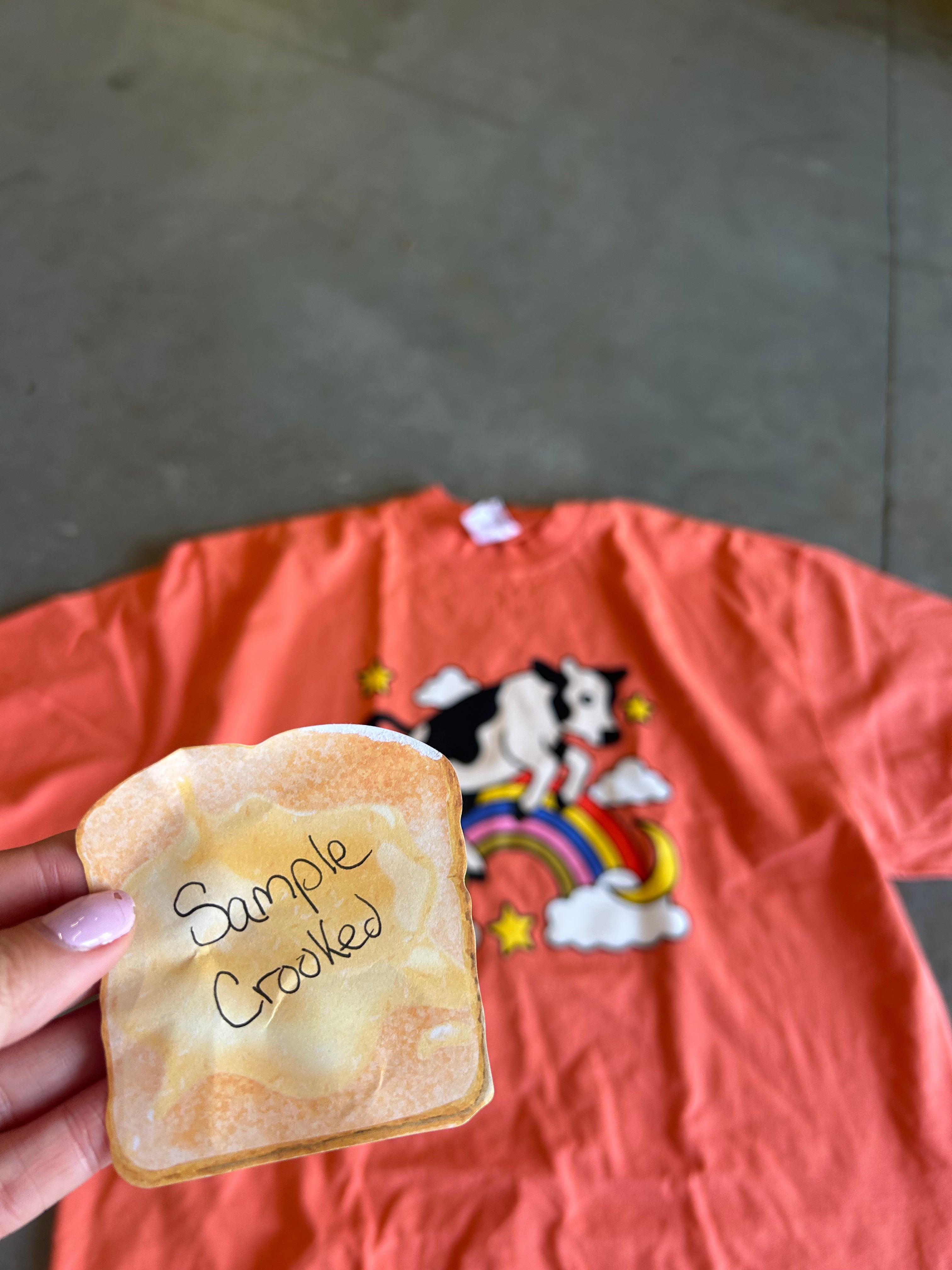 A hand holds a novelty toast prop with the words 'Sample Crooked' written on it, positioned in front of an orange graphic t-shirt laid flat on a concrete floor.