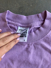 A close-up of a lavender purple t-shirt showing the inner neck label which reads 'Cruiser Weight Shirts' and 'L'. A hand with light pink manicured nails is pointing to the label.