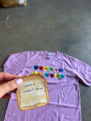 A hand holds a realistic-looking novelty toast prop in front of a lavender T-shirt laid flat on a concrete floor. The shirt has colorful letter tiles spelling 'SCHOOL IS COOL' and a small white tag near the collar.