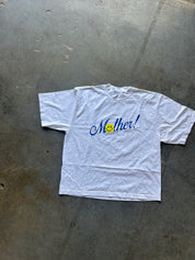 A white short-sleeve t-shirt with 'Mother!' written in blue cursive script and a yellow smiley face, laid flat on a gray concrete floor.