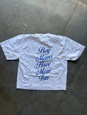 A white short-sleeve t-shirt laid flat on a gray concrete floor, featuring the phrase 'Boy Moms Have More Fun' printed in blue cursive text on the back.