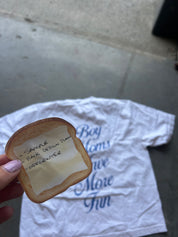 A hand holds a small, bread-shaped item with handwritten text on a piece of tape, positioned in front of a white graphic t-shirt on a concrete floor.