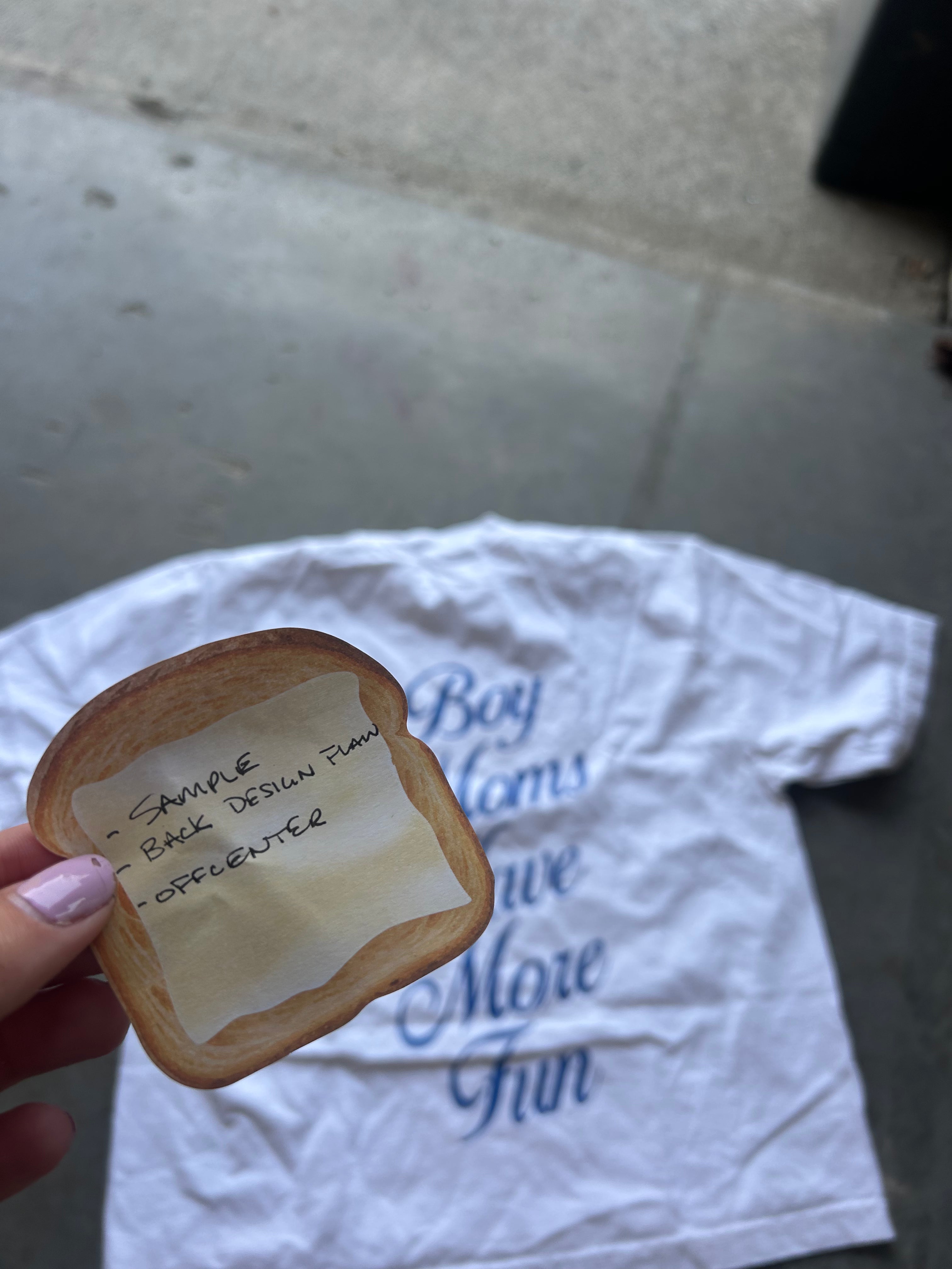 A hand holds a small, bread-shaped item with handwritten text on a piece of tape, positioned in front of a white graphic t-shirt on a concrete floor.