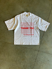 A white short-sleeve T-shirt laid flat on a concrete floor, featuring a red graphic print that reads 'THANK YOU for raising your funds' with repeated 'THANK YOU' text in red and pink.