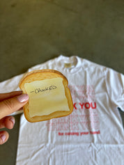 A hand holds a realistic plastic slice of toast with a yellow sticky note that says '-CROOKED' in front of a white graphic t-shirt on a concrete floor.