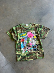A vintage short-sleeve t-shirt with a green and brown camouflage pattern, featuring a colorful retro graphic collage on the front with the text 'Go Angel' and various pop culture icons, laid flat on a concrete floor.