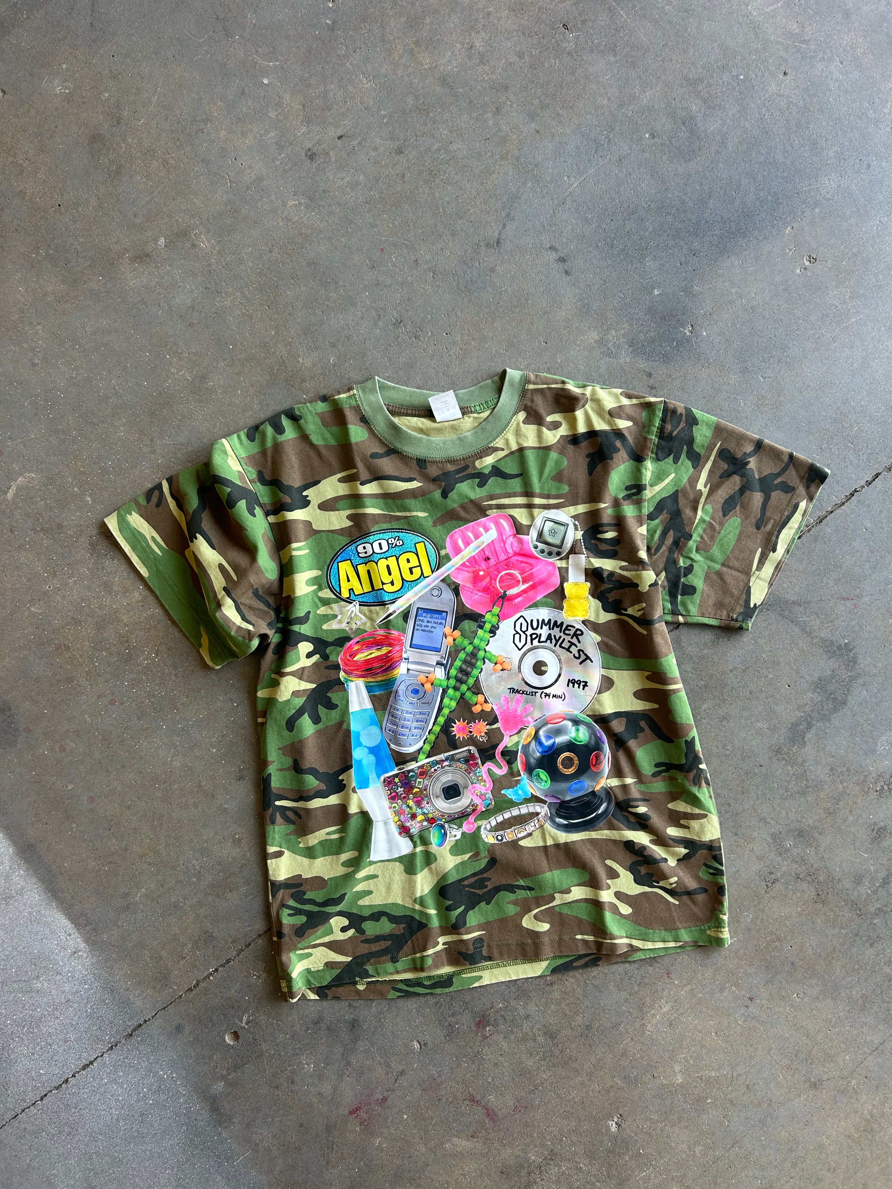 A vintage short-sleeve t-shirt with a green and brown camouflage pattern, featuring a colorful retro graphic collage on the front with the text 'Go Angel' and various pop culture icons, laid flat on a concrete floor.