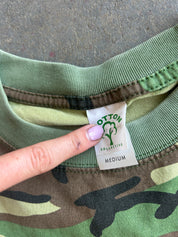 Close-up of a camouflage sweatshirt collar showing a finger pointing to the Cotton Collective brand label, with a gray concrete floor background.
