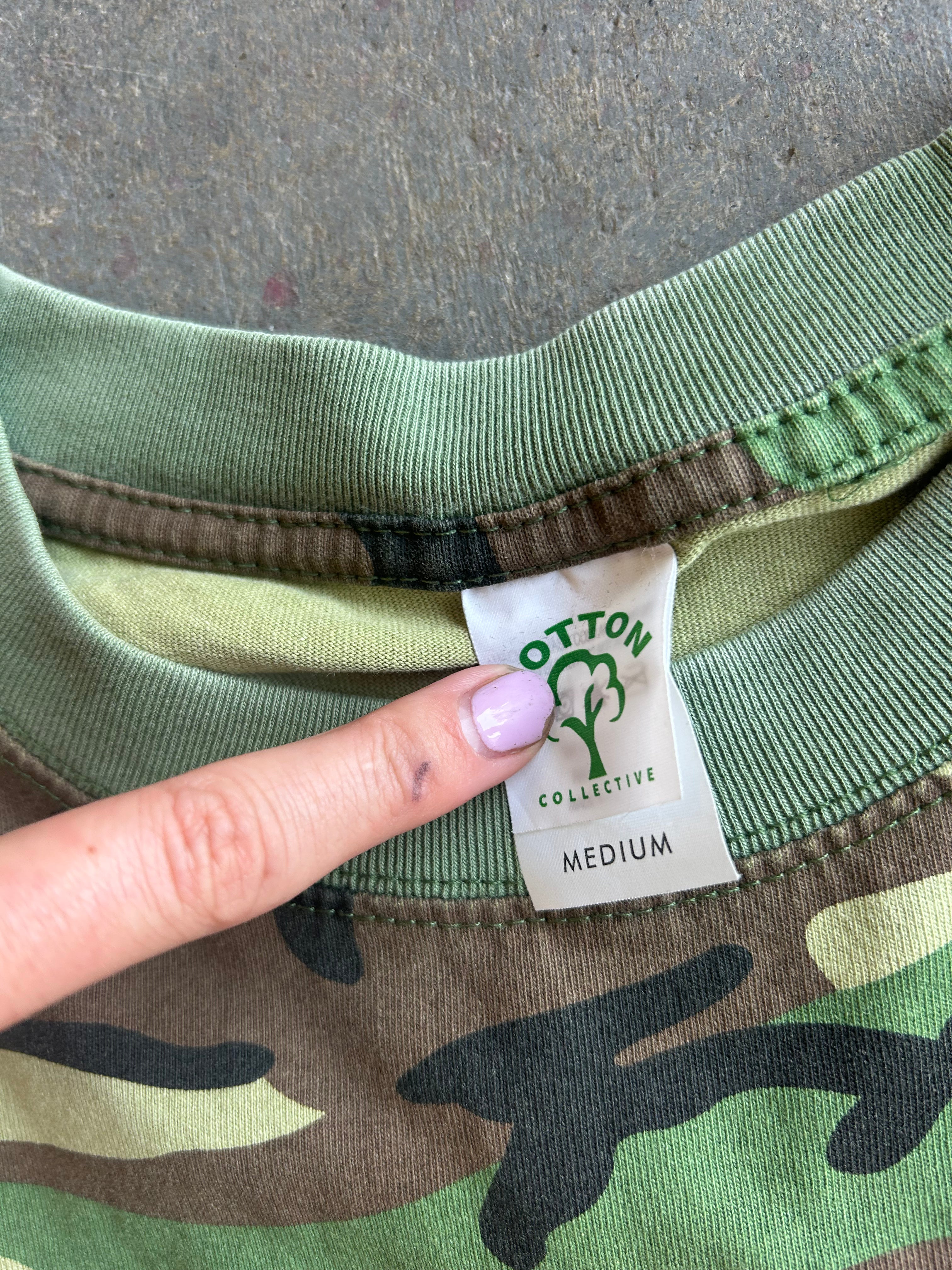Close-up of a camouflage sweatshirt collar showing a finger pointing to the Cotton Collective brand label, with a gray concrete floor background.