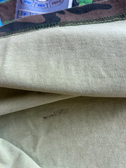 Close-up of a light beige or off-white knit fabric, showing the texture and a small dark stain, with a camouflage-patterned trim visible at the top edge.