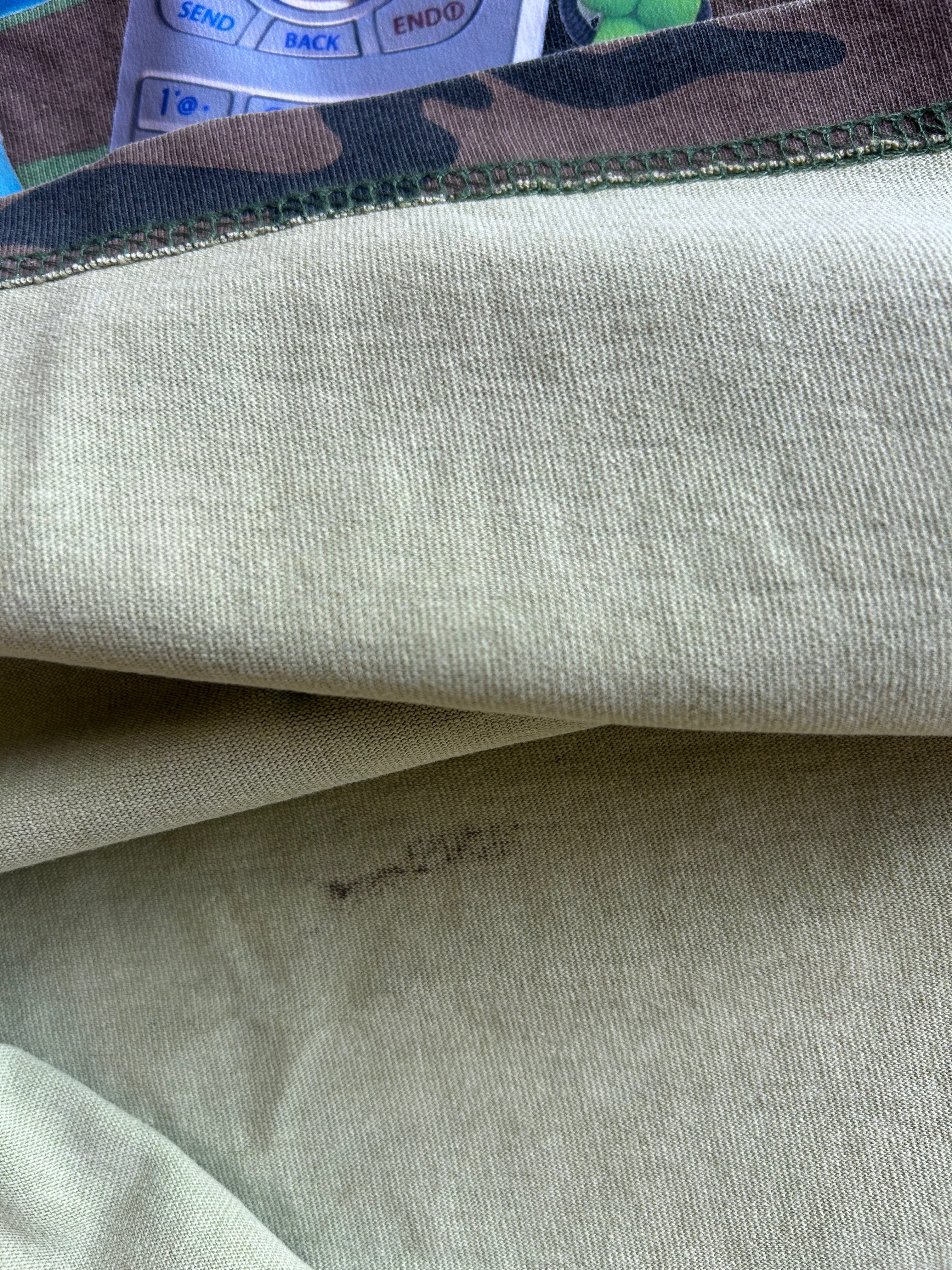 Close-up of a light beige or off-white knit fabric, showing the texture and a small dark stain, with a camouflage-patterned trim visible at the top edge.