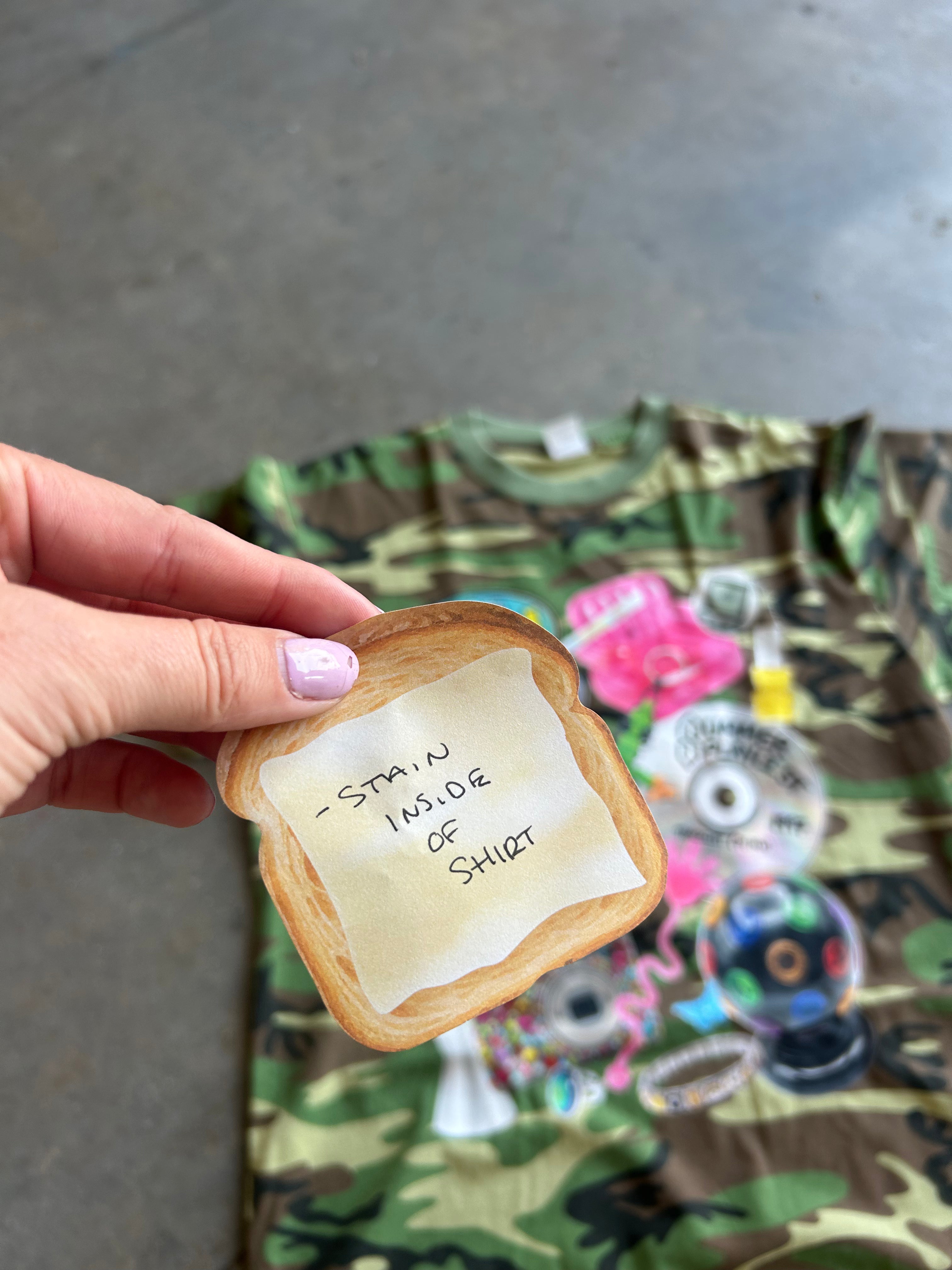 A hand holds a toasted bread slice-shaped sticker with handwritten text reading '- STAIN INSIDE OF SHIRT' over a camouflage graphic t-shirt laid on a concrete floor.