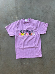 A light purple short-sleeve t-shirt with a graphic reading 'Til' Death we don't' in black and multicolor lettering, laid flat on a textured gray concrete surface.