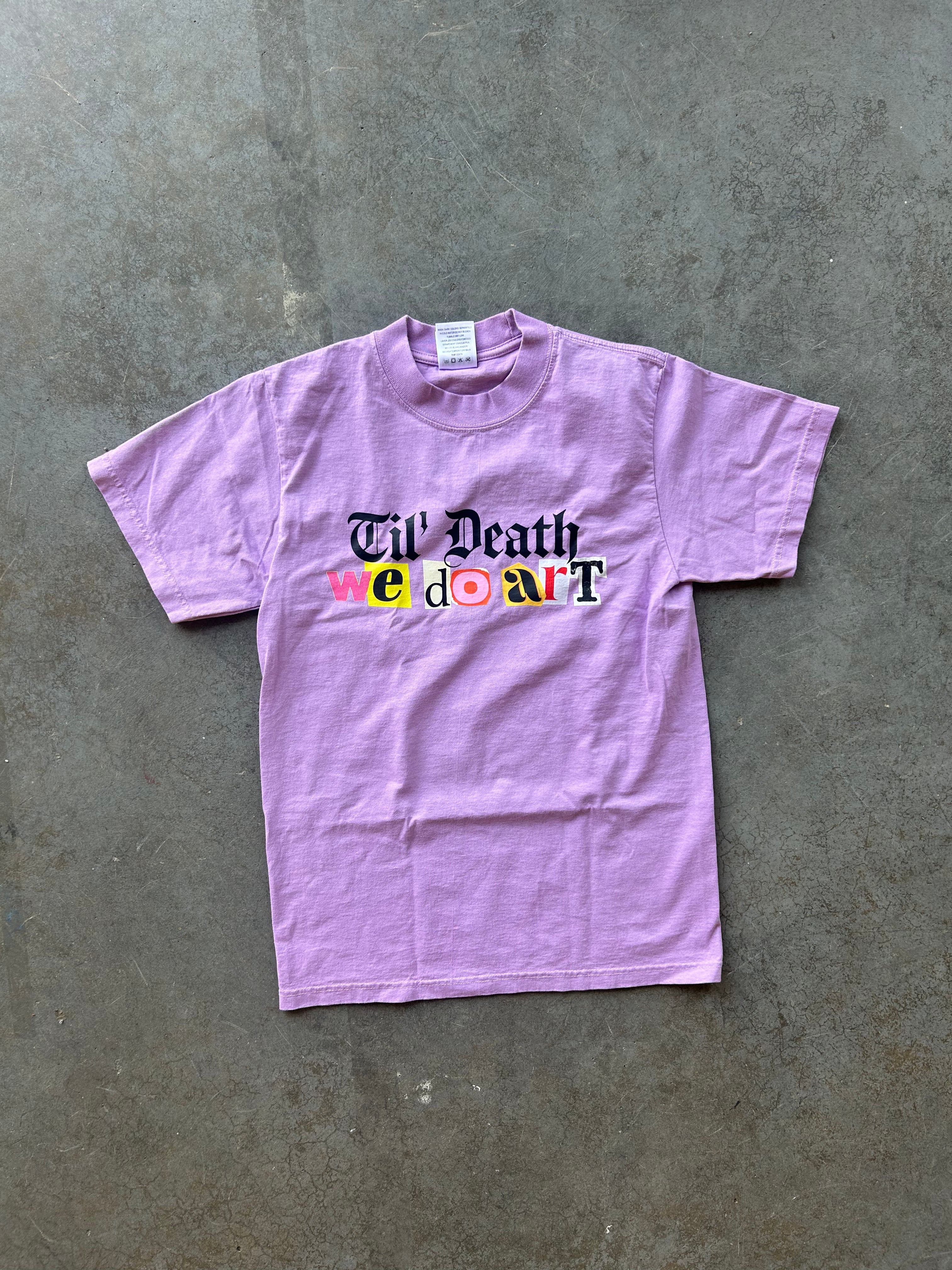 A light purple short-sleeve t-shirt with a graphic reading 'Til' Death we don't' in black and multicolor lettering, laid flat on a textured gray concrete surface.
