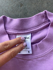 A close-up of a lavender purple t-shirt showing the inner neck label with a hand pointing to the label.