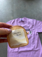 A hand holds a realistic foam slice of toasted bread with a handwritten note that says 'CROOKED PRINT'. In the blurred background, a light purple t-shirt is laid out on a gray floor.