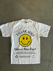 A white short-sleeve graphic t-shirt laid flat on a gray concrete floor. The shirt features a large yellow smiley face in the center with the words 'THANK YOU!' arched above it and 'Have a Nice Day!' printed below in black.