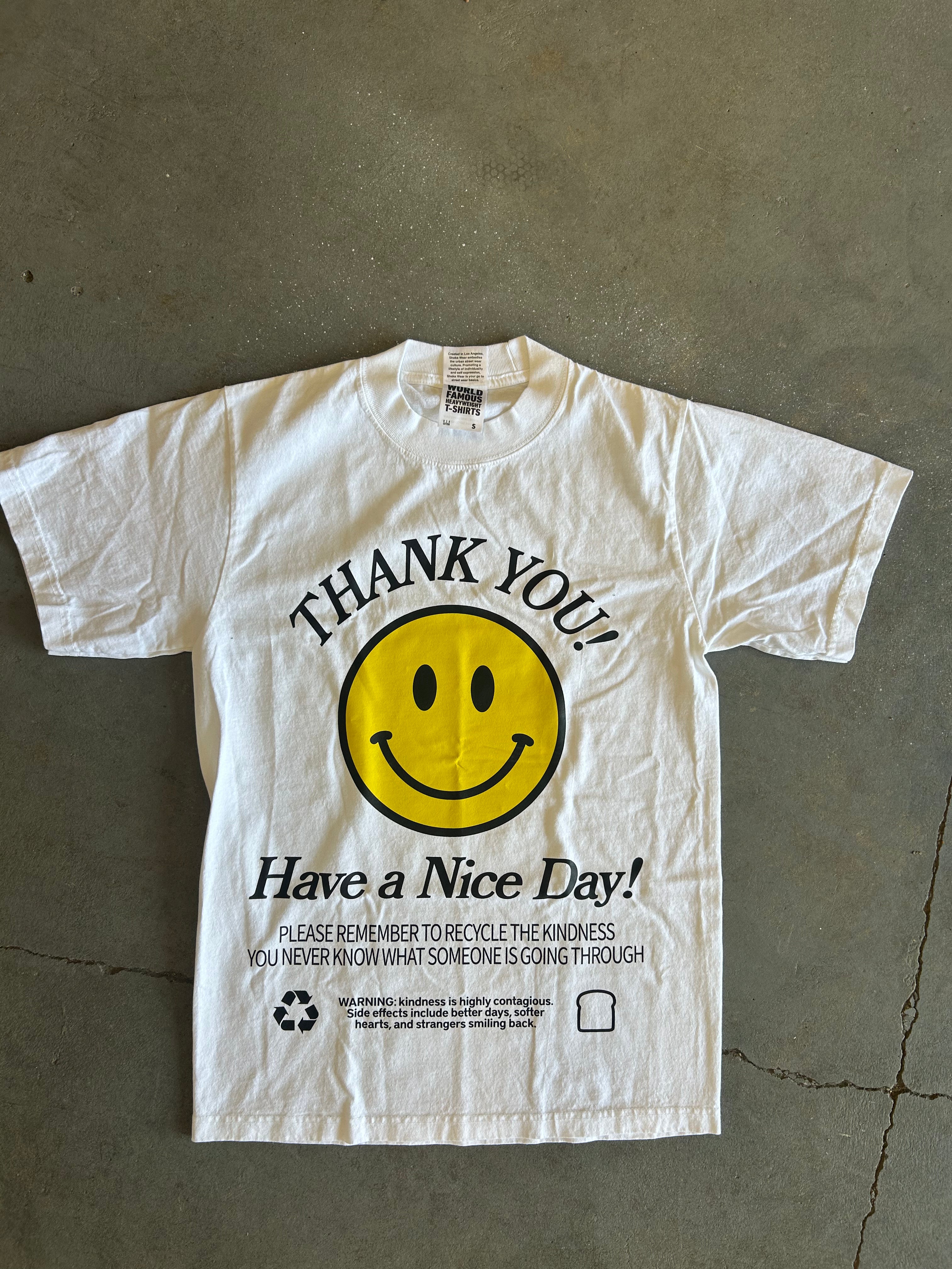A white short-sleeve graphic t-shirt laid flat on a gray concrete floor. The shirt features a large yellow smiley face in the center with the words 'THANK YOU!' arched above it and 'Have a Nice Day!' printed below in black.