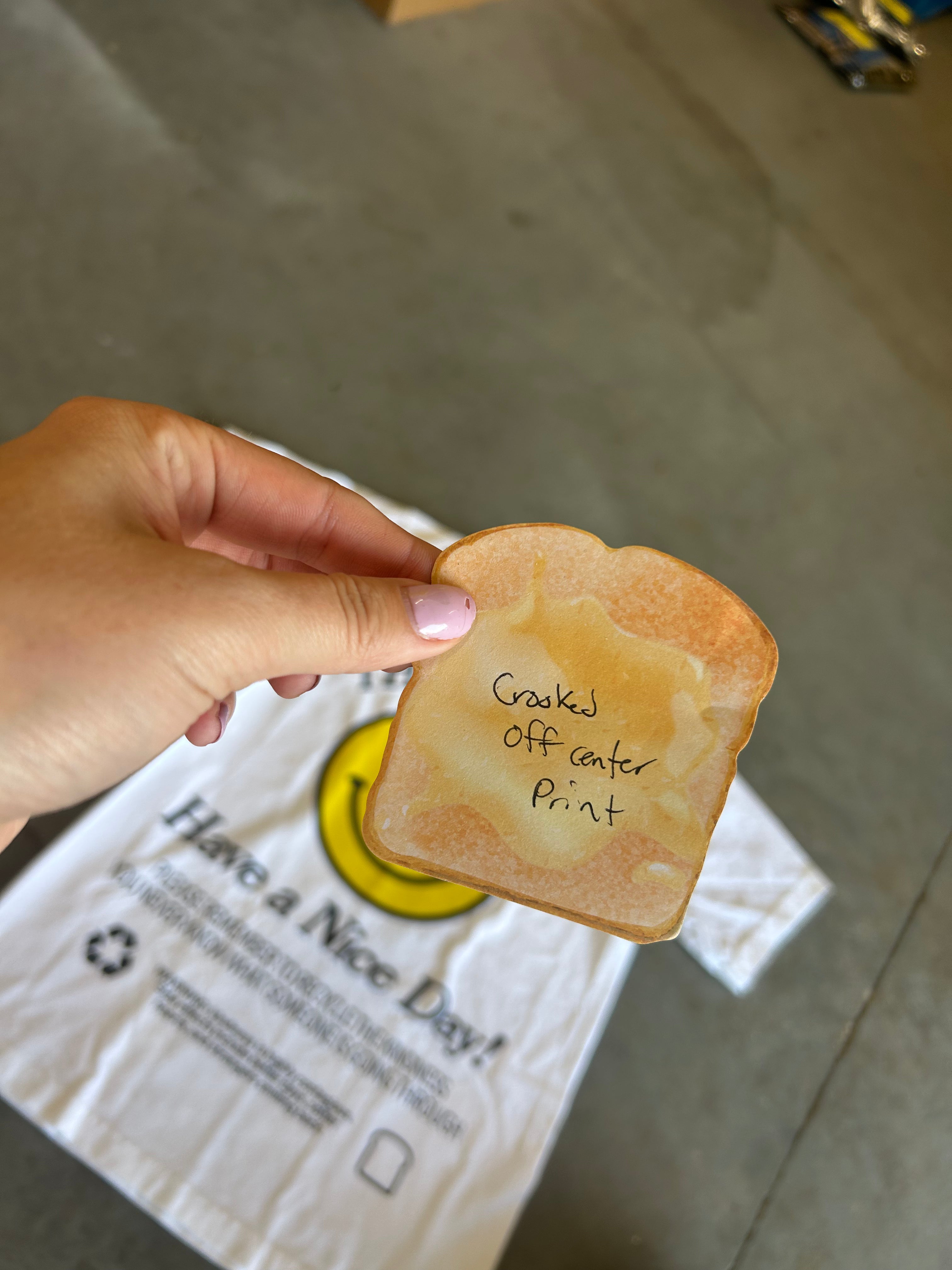 A hand holds a novelty slice of toast printed with the text 'Cracked off center Print' above a white tote bag with a yellow smiley face and the text 'Have a Nice Day!'.