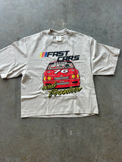 A light beige short-sleeve t-shirt laid flat on a gray concrete floor, featuring a graphic print of a red race car with the text 'FAST CARS and Freedom'.