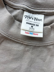 Close-up of a taupe-gray oversized t-shirt neck label by Shaka Wear, showing 'Streetwear Essentials Oversize Tee' and 'SuperMAX Heavy' text.